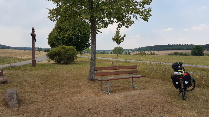 Rechts ein Fahrrad, mittig eine Sitzbank, daneben ein kleiner Baum im Hintergrund Landschaft und Wiese.