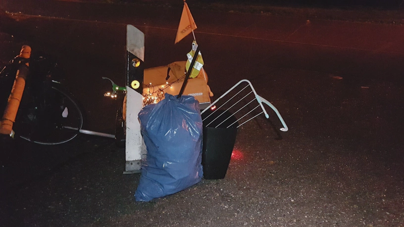 Anhänger mit dem gesammelten Müll, der davor gestellt wurde, und Teil des Fahrrads. Am Anhänger hängt eine eingeschaltete Lichterkette.