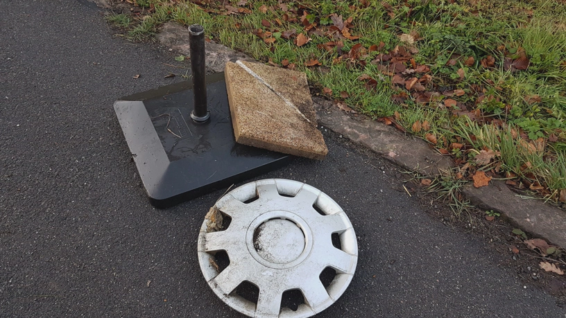 Reifenkappe eines Autos, Steinplatte und Standbein mit Gewicht für Sonnenschirm liegen am Straßenrand.