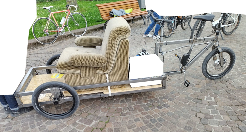 Ein Lastenrad mit LadeflÃ¤che vorne, auf dem ein Sessel mit Stoffbezug befestigt ist.