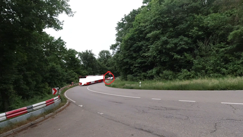 Ein Kurvenbereich einer Landstraße. Weiter hinten steht das rote Auto in der Kurve und wurde gerade von einem Busfahrer mit Bus überholt, ein Fahrer eines Autos überholt in diesem Moment.