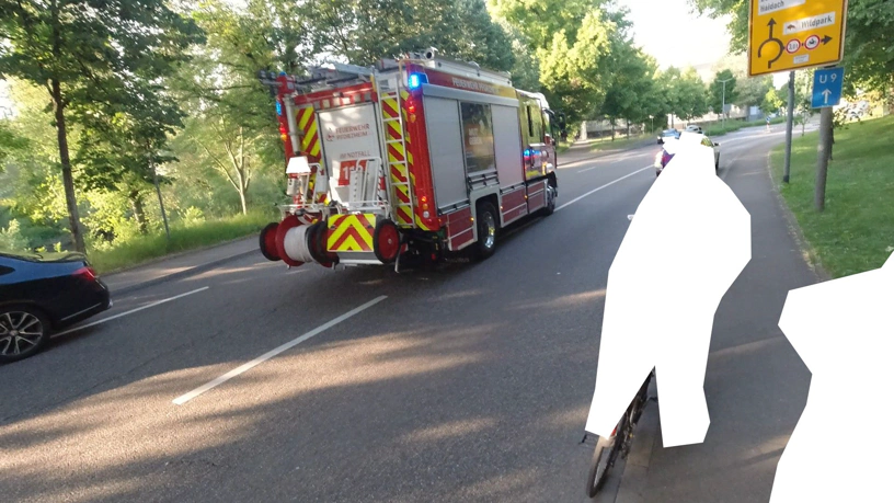 Feuerwehrfahrzeug mittig auf der StraÃŸe, links Teil eines stehenden Autos, rechts unkenntlich gemachte Radfahrende, die warten, eine davon auf dem Gehweg.