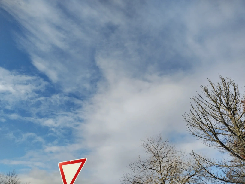 Blauer Himmel mit Schleierwolken. Unten links ein 'Vorfahrt gewähren'-Schild und rechts Teile eines Baums.